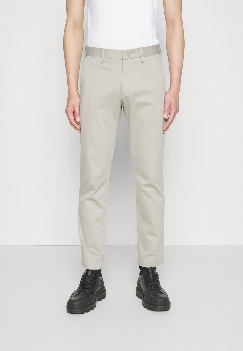 Man med ljusbeiga raka byxor med bältesöglor och svarta kraftiga snörskor, stående mot en vanlig vit bakgrund.