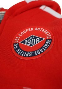 Le tissu du sweat-shirt rouge présente un badge circulaire avec le texte contrastant en bleu marine « Lee Cooper Authentic Heritage » et « EST 1908 ».