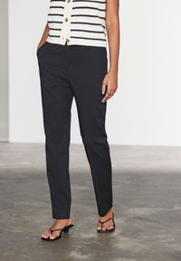 Svarta skräddarsydda byxor med smal passform och mjuk textur, med sidofickor och en slät framdesign, kombinerade med svarta flip-flops.