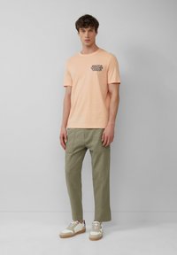 Perzikkleurig katoen T-shirt met korte mouwen, met zwarte grafische tekst op de linkerkant van de borst; gecombineerd met lichtgroene cargo-broek en witte sneakers.