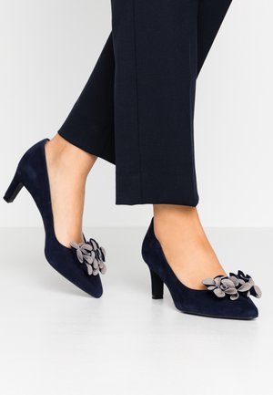 Pieds chaussés d'escarpins kitten en daim bleu marine avec des appliques florales grises, assortis à un pantalon court noir sur un fond uni.