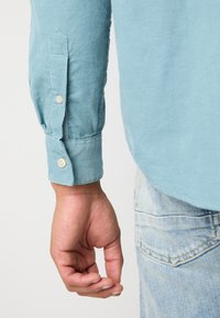 Mano rilassata di lato, indossa una camicia in velluto a coste blu chiaro con bottoni beige e il taschino dei jeans blu chiaro visibile.