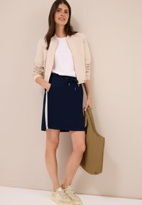 Kvinna som står mot en beige vägg, klädd i beige jacka, vit skjorta, marinblå kjol med vit rand, beige sneakers, och håller en brun tote-väska.