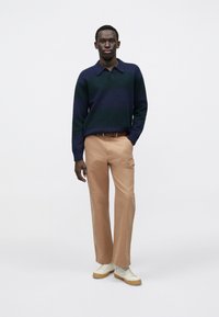 Tengerészkék és zöld csíkos kötött polo ing, bézs nadrág, fehér sportcipő bézs talppal, egyszerű fehér háttér előtt állva.