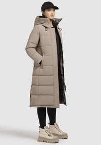 Beige gewatteerde lange jas met een capuchon, voorzien van een zip-up voorkant en zijzakken. Draagt bij beige chunky-sole laarzen en zwarte legging.