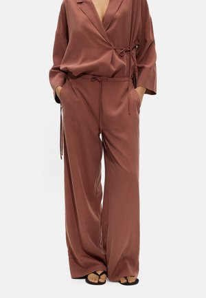 Kobieta w luźnej brązowej satynowej bluzce typu wrap, szerokich spodniach i czarnych sandałach, ręce w kieszeniach, na białym tle.
