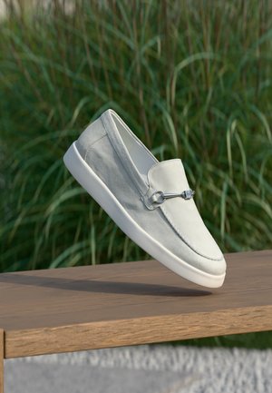 Hellgrauer Wildleder-Slipper mit weißer Sohle und Metallschnalle, schwebend in der Luft über einer Holzoberfläche mit grünem Gras im Hintergrund.