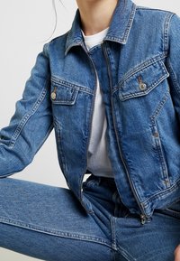 Denimová bunda v světle modré barvě, se dvěma náprsními kapsami se stříbrným kováním a zipovým zapínáním. Tkanina má hladký povrch.