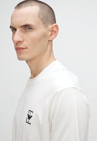 Balta ilgomis rankovėmis marškinėliai su juodu logo kairėje krūtinės pusėje; minkšta medžiaga, apvali iškirptė ir laisvas fasonas.