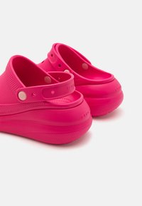 Un par de Crocs de color rosa brillante con correas en el talón, suelas texturizadas y agujeros de ventilación sobre un fondo blanco.