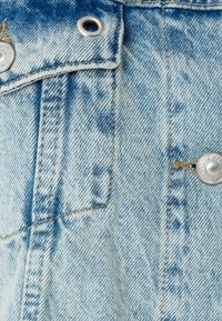 Ljusblå denimtyg med ett urblekt mönster, med silverfärgad metall och synliga sömnadsdetaljer, inklusive en framficka och knapp.