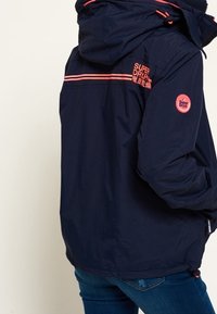 Marine vandtæt jakke med hætte, pink logo og stribede detaljer. Har elastiske manchetter og en snor i kanten, lavet af et letvægtsmateriale.