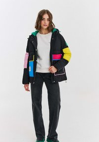 Žena oblečená v čiernej púfnej bunde s farebnými nášivkami, bielej mikine, čiernych džínsoch a zelených topánkach, stojaca proti bielej stene.