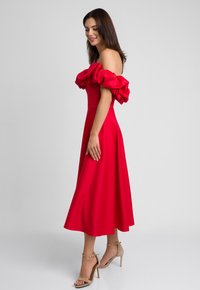 Czerwona sukienka bez ramiączek z dużymi falbaniastymi rękawami, dopasowanym gorsetem i zwiewnym spódnicą w kształcie litery A. Noszona z beżowymi sandałami na obcasie.