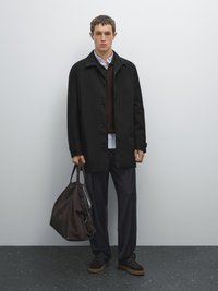 Schwarzer Knopfverschlussmantel über einem braunen Pullover, einem weißen Hemd mit Kragen, dunkelblauen Hosen und dunkelbraunen Schuhen. Trägt eine braune Tasche.