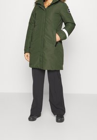 Parka aislante verde con capucha forrada de piel, diseño acolchado vertical, bolsillos laterales y acentos blancos en las mangas. Combinada con pantalones y zapatos negros.