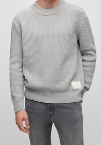 Pull en maille grise avec une texture côtelée et un col rond. Présente une petite étiquette blanche sur le côté inférieur gauche. Porté avec un jean gris.