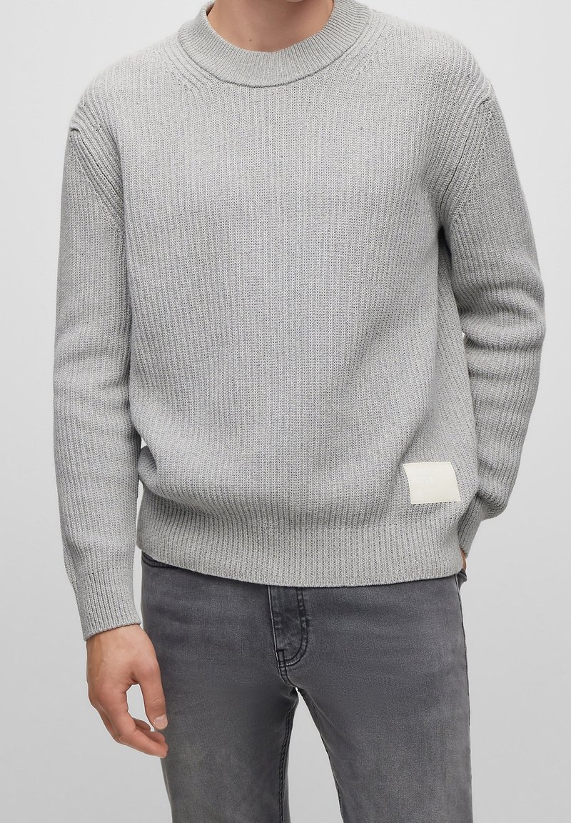 Pull en maille grise avec une texture côtelée et un col rond. Présente une petite étiquette blanche sur le côté inférieur gauche. Porté avec un jean gris.