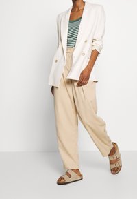 Vit dubbelknäppt kavaj, grön och vit randig t-shirt, beige veckade byxor, beige sandaler med två spännen, naturlig textur.