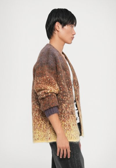 Kootud kardigan soojades pruunides, oranžides ja kollastes toonides, tekstuurse mustriga, ülekaalulise lõikega ja triibuliste varsikutega.