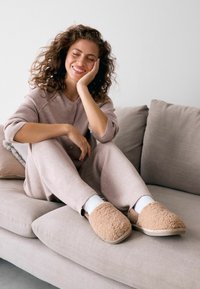 Beige, flauschige Hausschuhe aus einem weichen Material mit runder Form. Getragen mit weißen Socken, kombiniert mit einem hellrosa, plüschigen Outfit auf einem grauen Sofa.