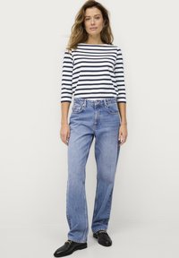 Gestreepte longsleeve shirt in marineblauw en wit, gecombineerd met lichtblauwe, iets baggy jeans en zwarte loafers met gouden accenten.