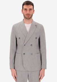 Uomo che indossa un giubbotto grigio a doppio petto con righe sottili, pantaloni abbinati e una t-shirt bianca girocollo, in posizione eretta.
