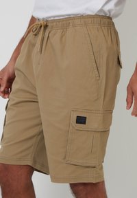 Beige cargoshorts med elastisk linning, snøre, sidelommer og en lomme på låret med et mærke. Holdbart stof.