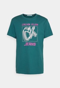 Turkusowa bawełniana koszulka z grafiką, na której widnieje "CALVIN KLEIN" w różowym kolorze oraz "JEANS" w pogrubionej czcionce na dole; krótki rękaw, okrągły dekolt.