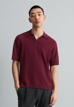 Jeune homme aux cheveux noirs courts, portant un pull bordeaux à manches courtes avec col et un pantalon noir, debout devant un fond gris.