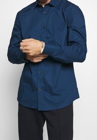 Camisa abotonada azul marino en una tela suave, con dobladillo curvado y cuello estándar. Los botones negros añaden detalle al tapete frontal.