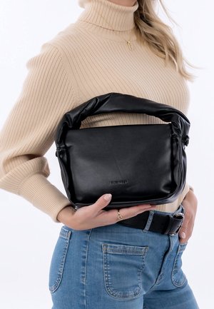 Femme en col roulé beige et jean bleu tenant un sac à main en cuir souple noir avec une anse rembourrée.