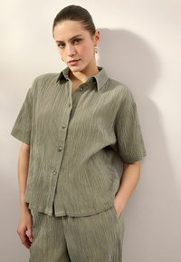 Licht olijfgroen overhemd met korte mouwen, met tekstuurrimpelingen door het hele shirt en een relaxte pasvorm. Gecombineerd met bijpassende onderkleding.