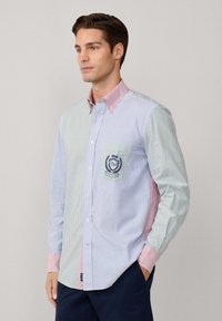 Hombre con camisa de botones a rayas de colores, con logo bordado en el bolsillo, pantalones navy y manos en los bolsillos, mirando hacia la izquierda contra un fondo liso.