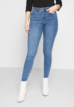 Vrouw gekleed in high-waist blauwe skinny jeans, zwart top met lange mouwen en witte enkellaarsjes, staand met één hand in haar zak.