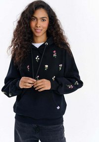 Černá mikina s kapucí s květinovou výšivkou v růžové a bílé barvě. Má přední kapsu, kapuci na stažení a žebrované manžety. Kombinováno s tmavými džíny.