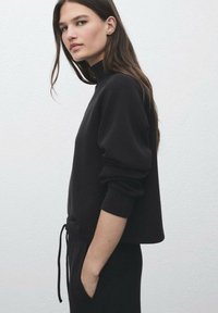 Sweatshirt noir court avec manches longues et col haut, présentant un détail à cordon au niveau de la taille. Texture lisse et design minimaliste.