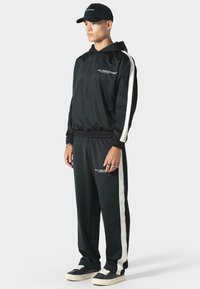 Tuta nera con strisce bianche ai lati; presenta una giacca con cappuccio e pantaloni con vita elastica. Indossato con un cappellino nero e sneakers.