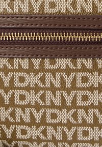 Tessuto testurizzato con un motivo ripetitivo "KNYD" in crema su uno sfondo oliva. Cerniera marrone con dettagli in metallo dorato.