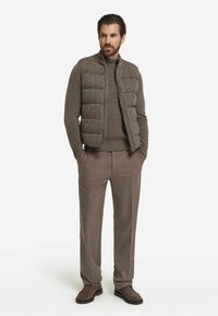 Braune gesteppte Weste über einem strukturierten Pullover, kombiniert mit beigen Hosen und braunen Schuhen. Glatter Stoff, klare Linien und neutrale Farbtöne.