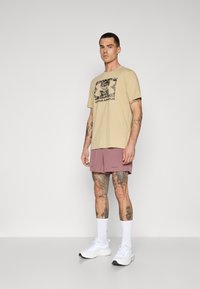 Beiges Baumwoll-T-Shirt mit schwarzem Under Armour-Logo, kombiniert mit helllila Shorts. Das Modell trägt weiße Schuhe und kniehohe weiße Socken.