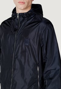 Giacca in nylon blu navy con zip frontale, cappuccio regolabile con cordino e una patch brandizzata sul petto. Texture liscia, design leggero.