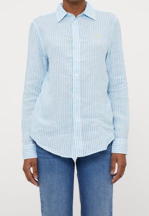 Person iført en lyseblå og hvid lodret striped skjorte med lange ærmer og blå jeans.