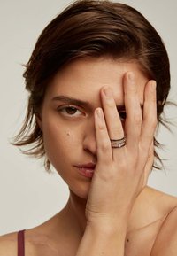 El anillo presenta tres bandas entrelazadas en tonos plateados y negros, mostrando una textura suave y un diseño minimalista. La modelo cubre un ojo con una mano.