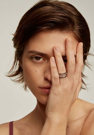 El anillo presenta tres bandas entrelazadas en tonos plateados y negros, mostrando una textura suave y un diseño minimalista. La modelo cubre un ojo con una mano.