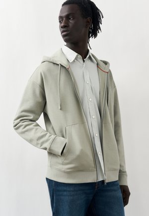 Fiatal sötét bőrű férfi dreadlockkal világosszürke cipzáras kapucnis pulóvert visel fehér gombos ing és kék farmer fölött, oldalra nézve.