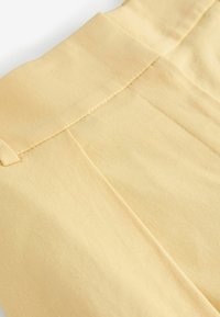 Housut pehmeästä keltaisesta kankaasta, jossa on sileä pinta, laskostettu etuosa ja siisti ommel vyötärön ympärillä.