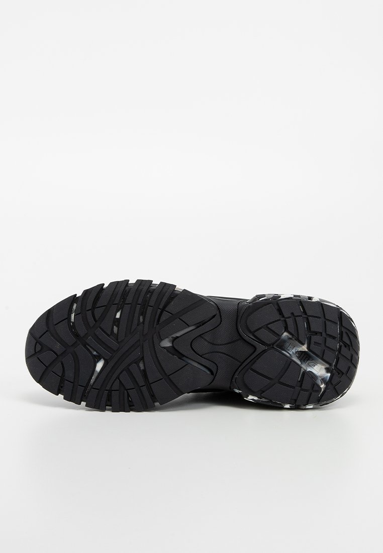 Semelle de chaussure de sport noire avec des rainures et des crans profonds, présentant une surface lisse et texturée ainsi que des éléments de rembourrage clairs.