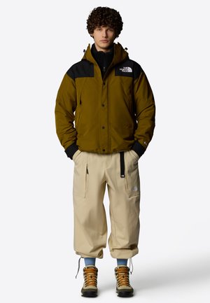 Oliwkowa zielona i czarna kurtka ocieplana, beżowe spodnie cargo, brązowe buty trekkingowe, w pozycji stojącej. Ubrania posiadają zamki błyskawiczne i logo.