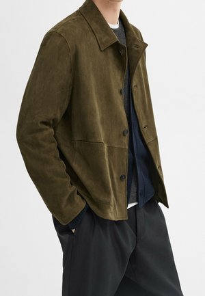Homme portant une veste en daim vert olive par-dessus un pull foncé et un pantalon noir, les mains dans les poches, sur un fond clair uni.